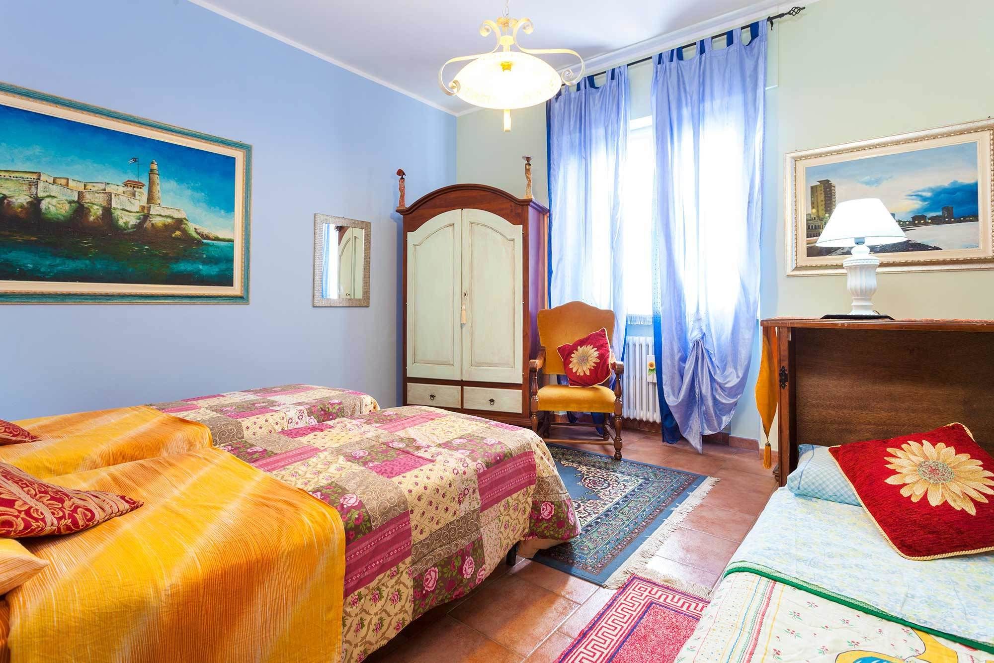 A bedroom with two beds, a wooden wardrobe, a chair with a cushion, and a painting on the wall.