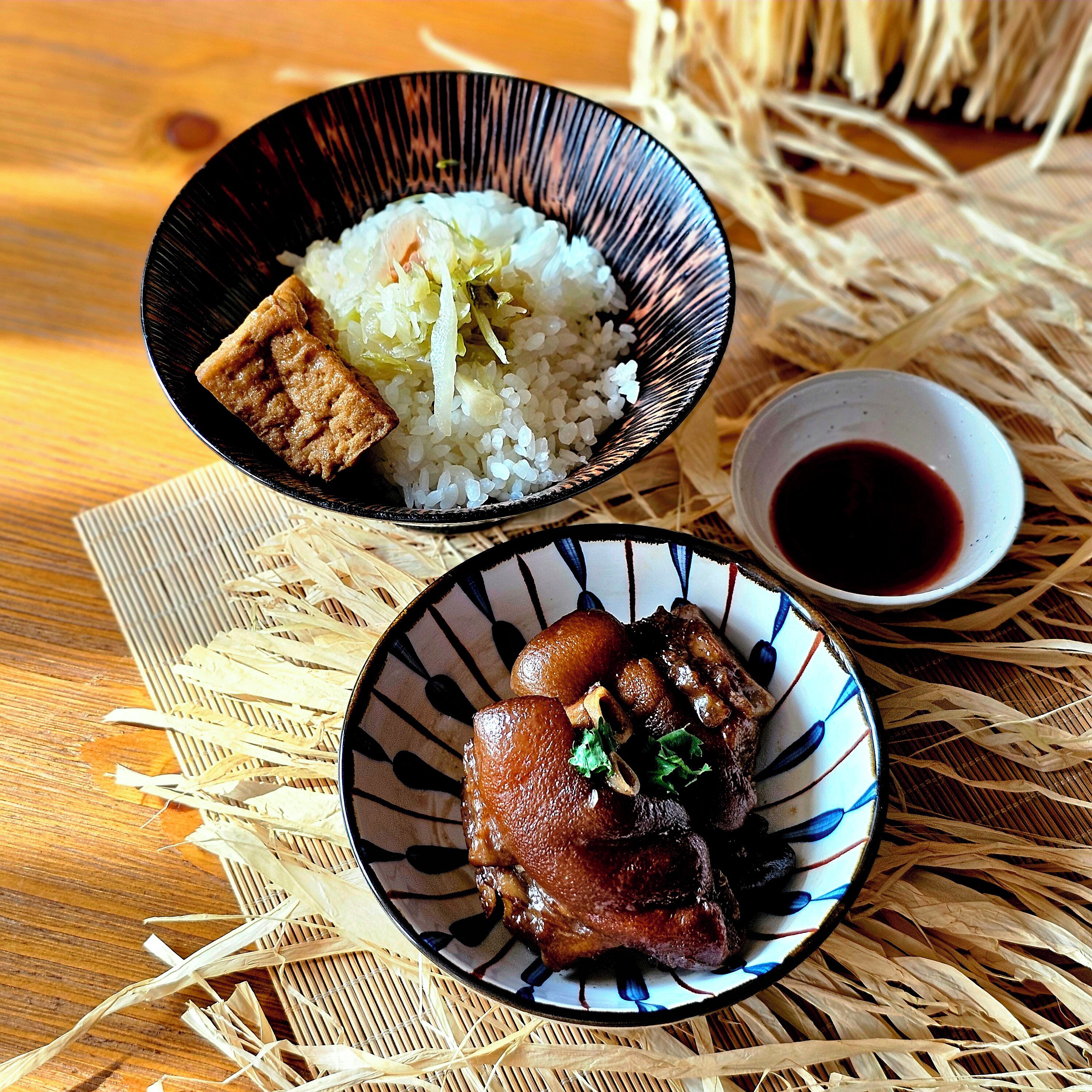 Une table en bois sur laquelle reposent deux bols de nourriture : l’un contient du riz et de la viande, et l’autre, un autre plat de viande. Les deux bols sont posés sur un tapis tissé.