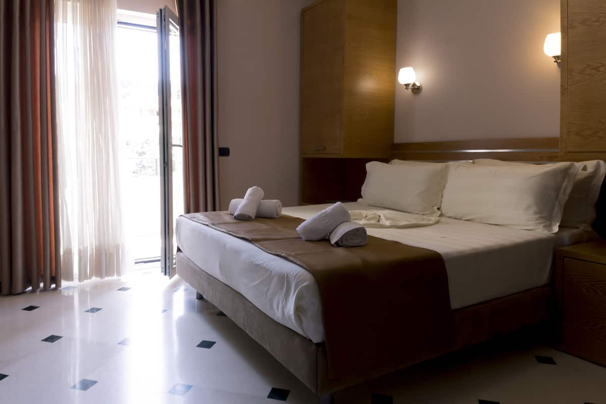 Una habitación de hotel con cama, mesita de noche, lámpara de pared y ventana con cortinas.