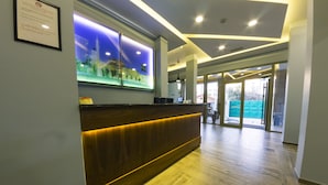 A reception area with a wooden reception desk, a digital display, and a view of the outside. - Hotel Pandora Residence (Tirana)
