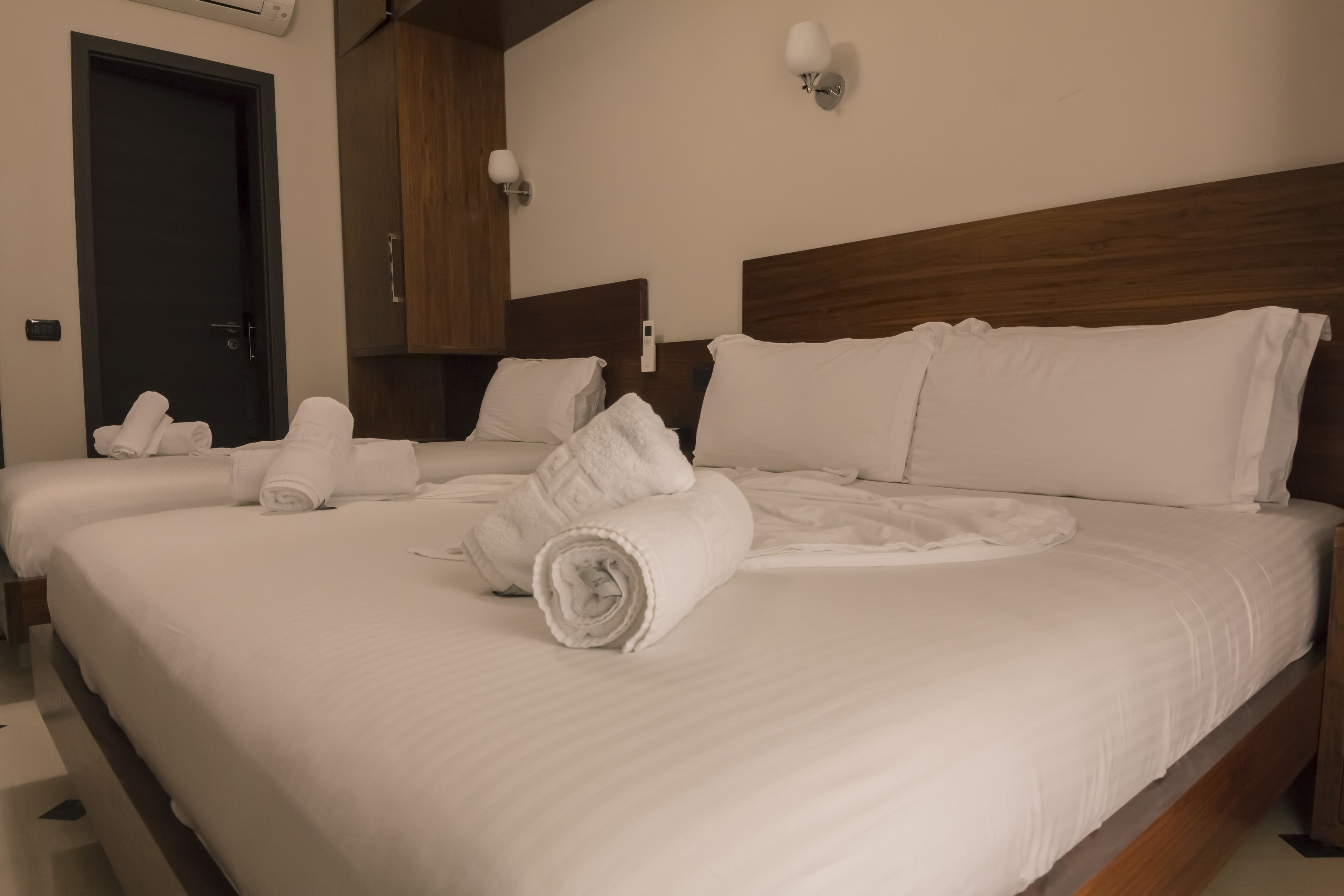 Habitación de hotel con dos camas, cada una con ropa blanca y toallas. Se observa un cabecero de madera, una lámpara de pared y una puerta con manija.