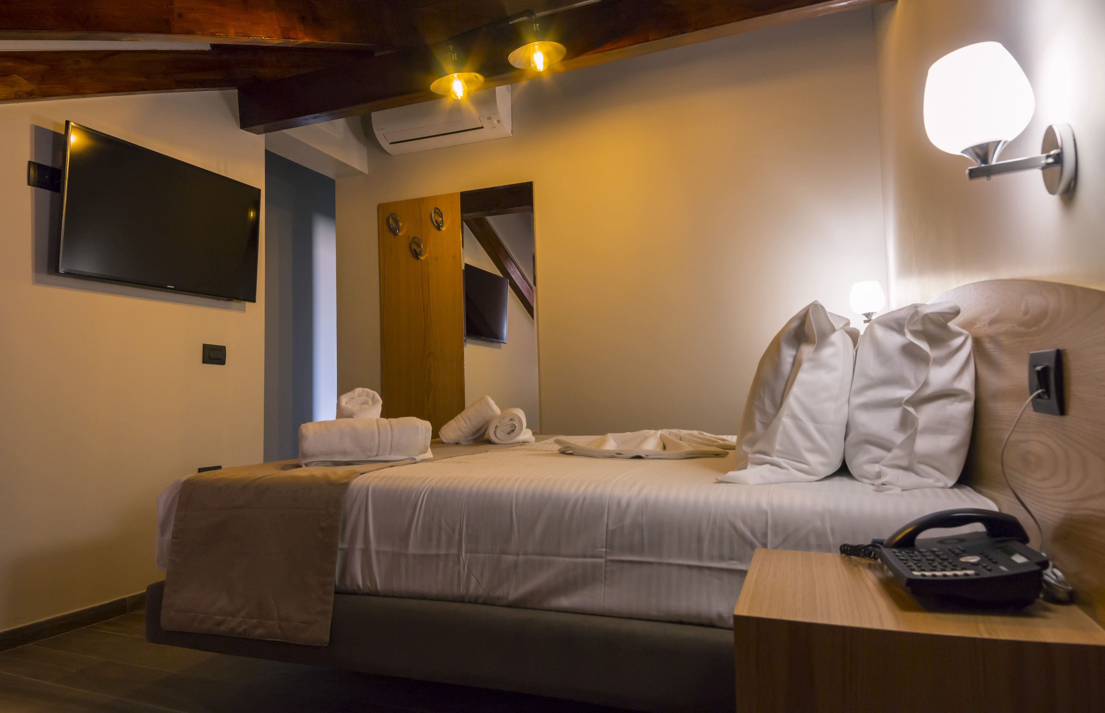 Habitación de hotel con cama, mesita de noche con teléfono, televisor montado en la pared y espejo.