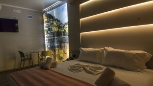 A hotel room with a large bed, a bedside table, a chair, and a view of a cityscape through a window. - Hotel Pandora Residence (Tirana)