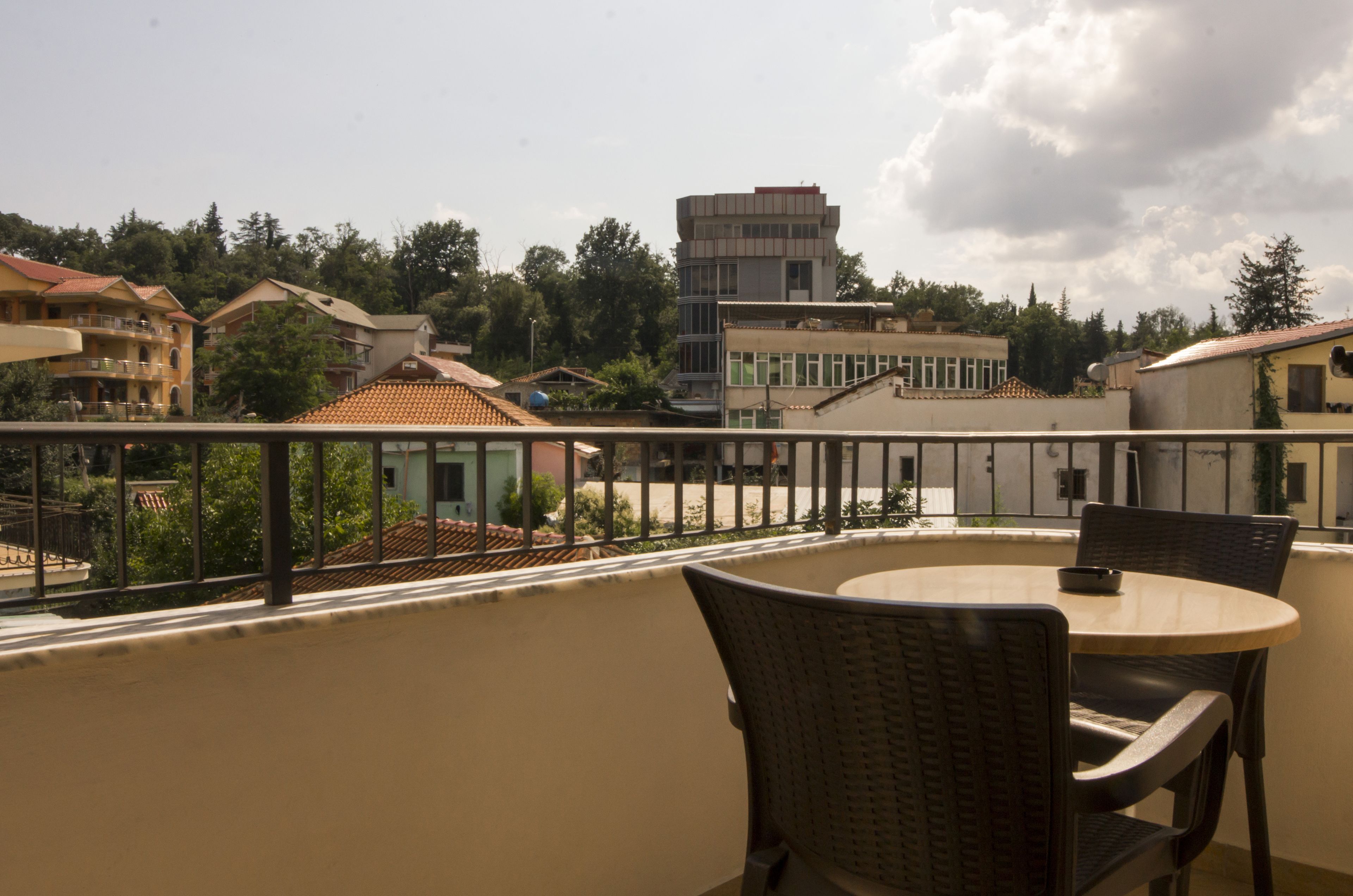 A balcony with wicker chairs and a table, overlooking a residential area with houses and a tall building.