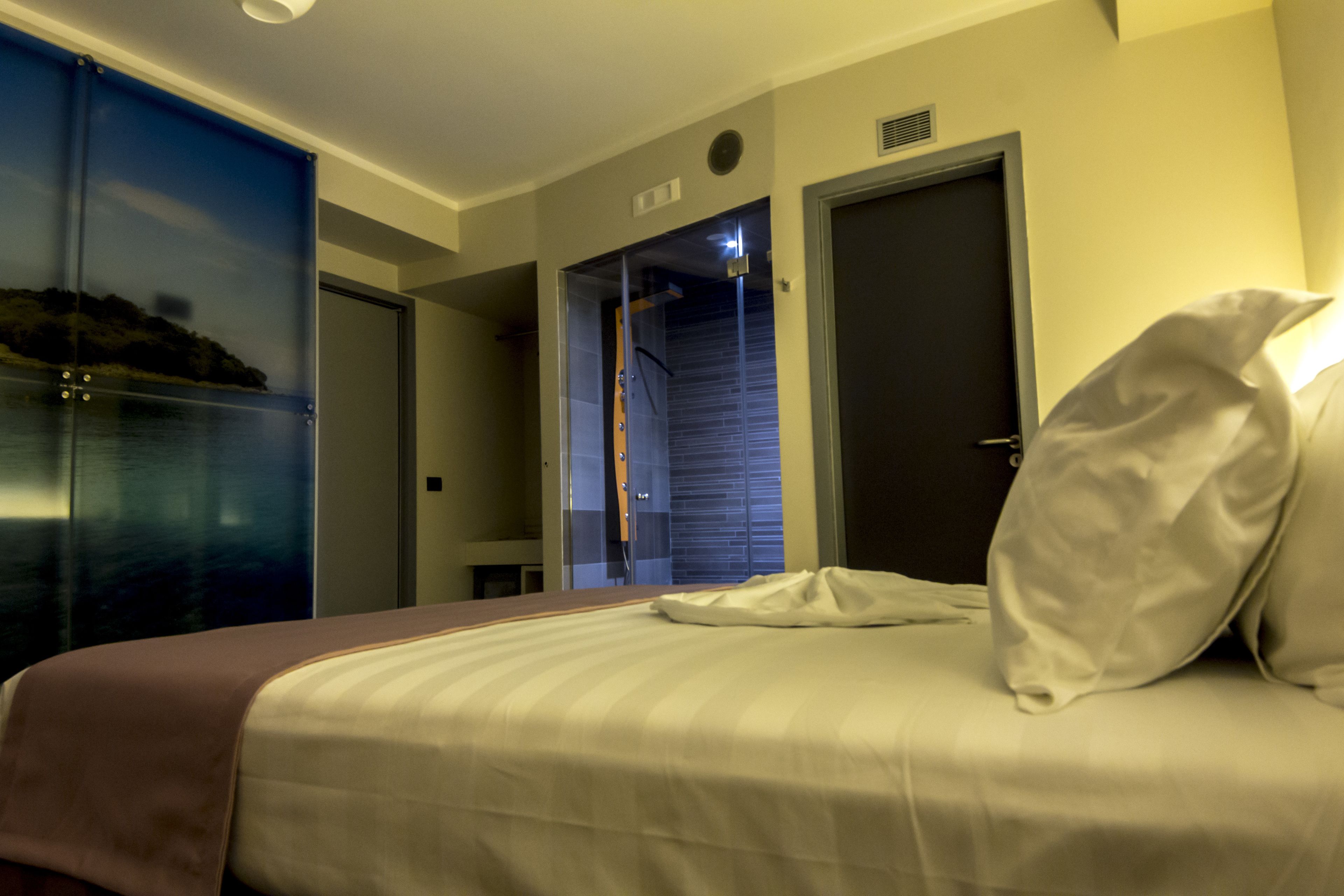 Ett hotellrum med en säng, ett nattduksbord och utsikt över en stadssiluett genom ett stort fönster.