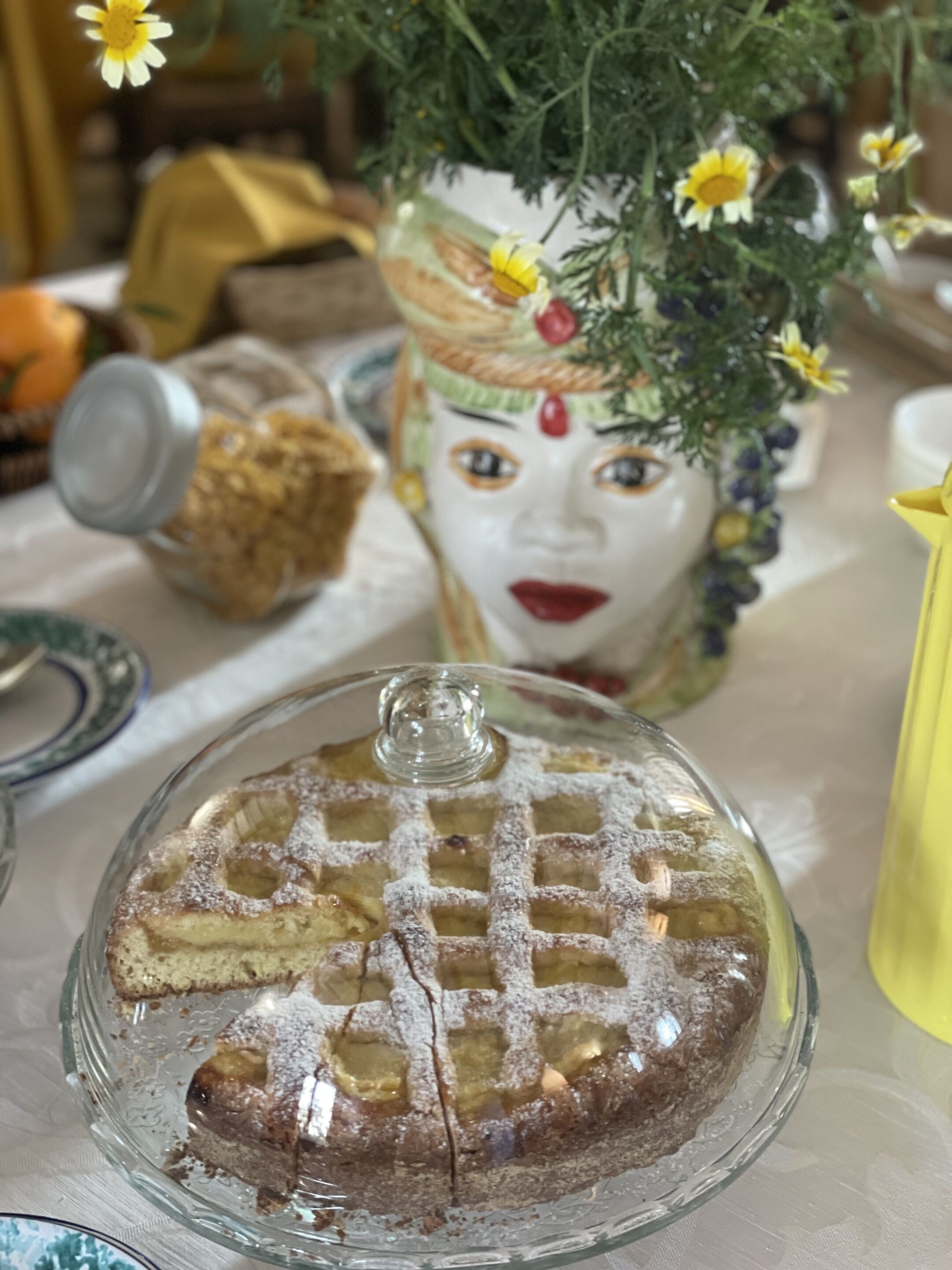 Ein Dessert auf einer Glasschale mit Gittermuster, eine dekorative Vase mit Blumen und ein gelbes Objekt auf dem Tisch.