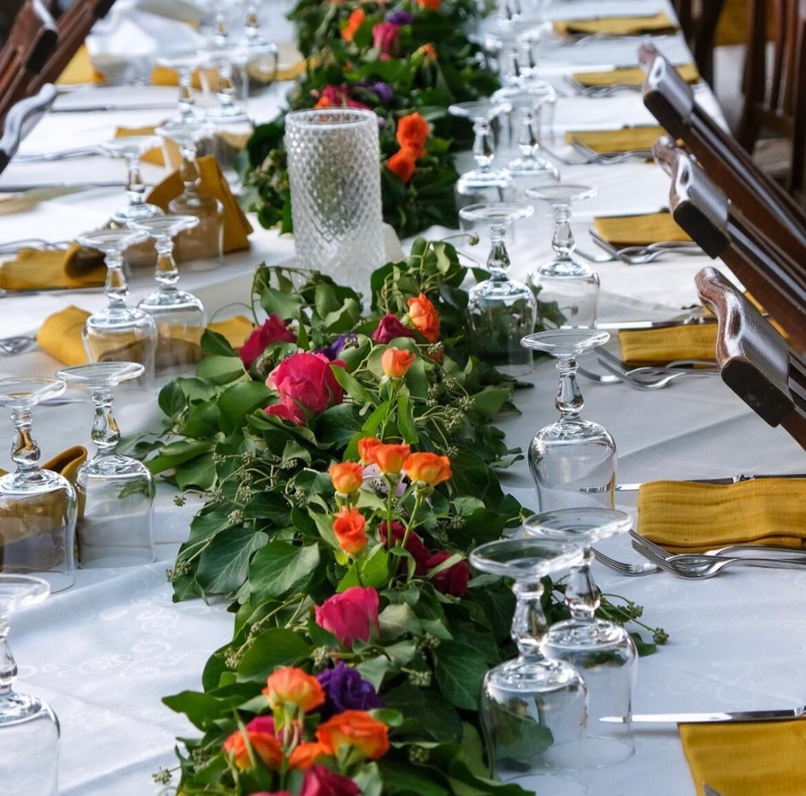 Mesa longa posta com taças, guardanapos e um arranjo floral.