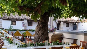 An outdoor dining area with tables set for a meal, surrounded by stone walls and a large tree. - Fattoria Manostalla (Partinico)