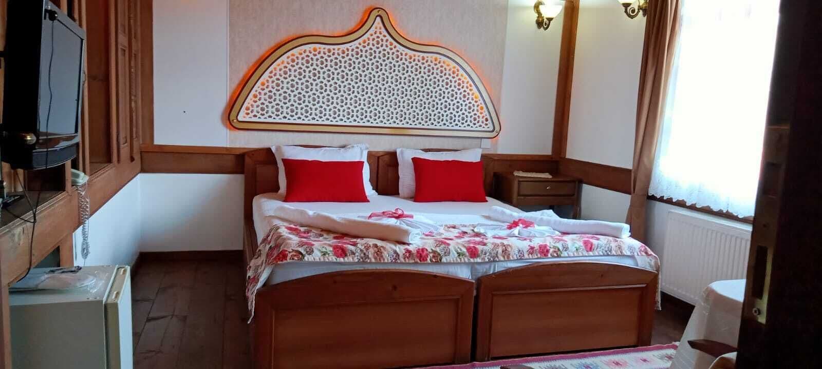 Une chambre d’hôtel avec un lit en bois, des oreillers rouges, une couvre-lit à motifs floraux, une télévision et une fenêtre avec des rideaux.