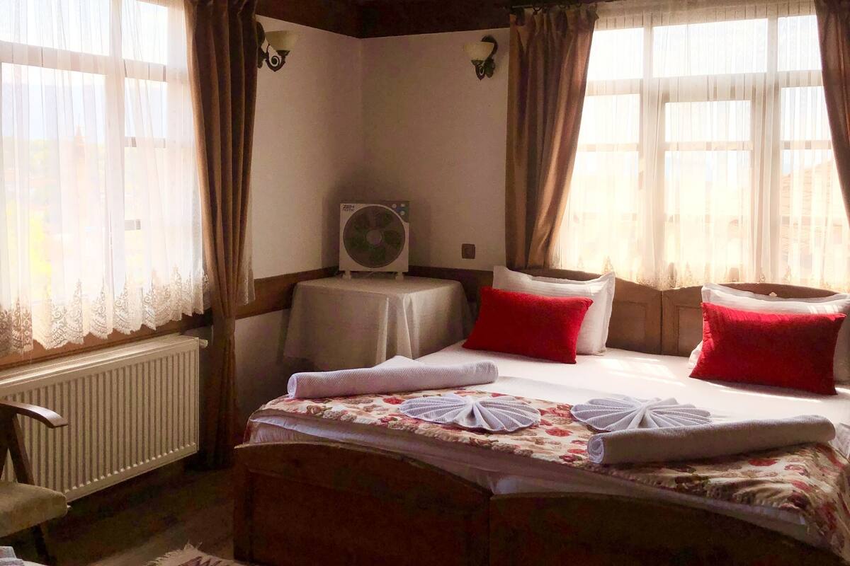 A room with a wooden ceiling, a bed with red pillows, a table with a fan, and a radiator.