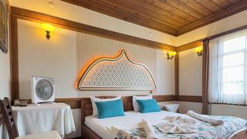 Een slaapkamer met een houten plafond, een groot bed met een bloemenbedekking, twee bedlampen, een wandheater en een tafeltje met een ventilator.
