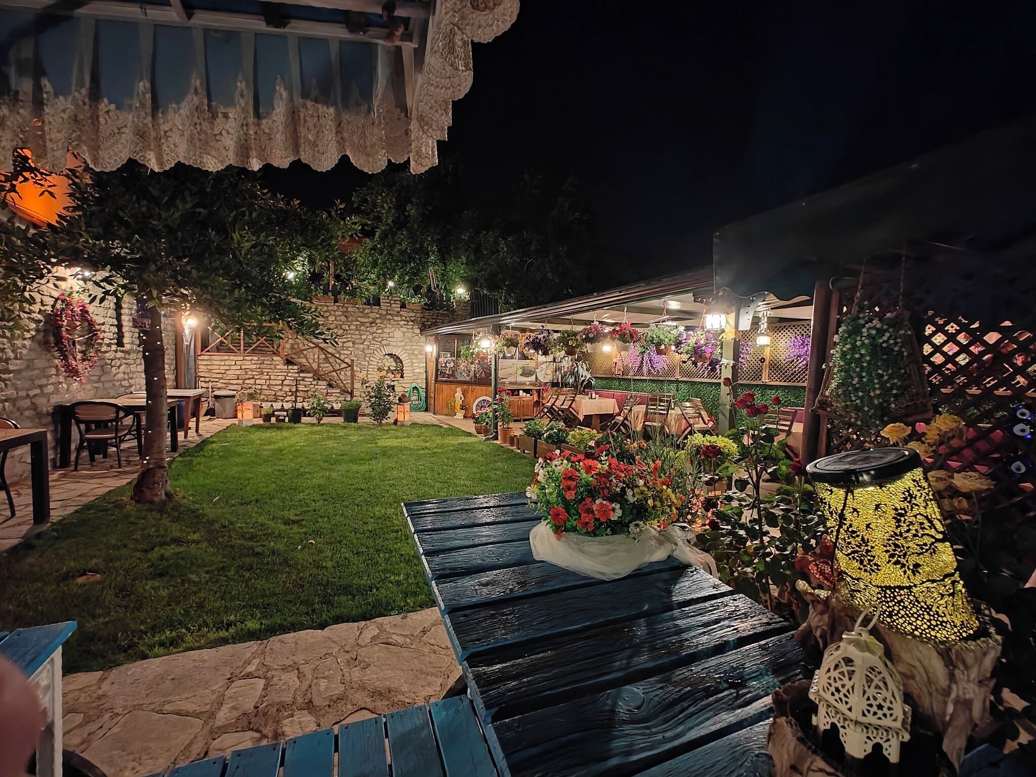 Un'area patio con un muro in pietra, tavoli e sedie in legno, circondata da piante e fiori in vaso.