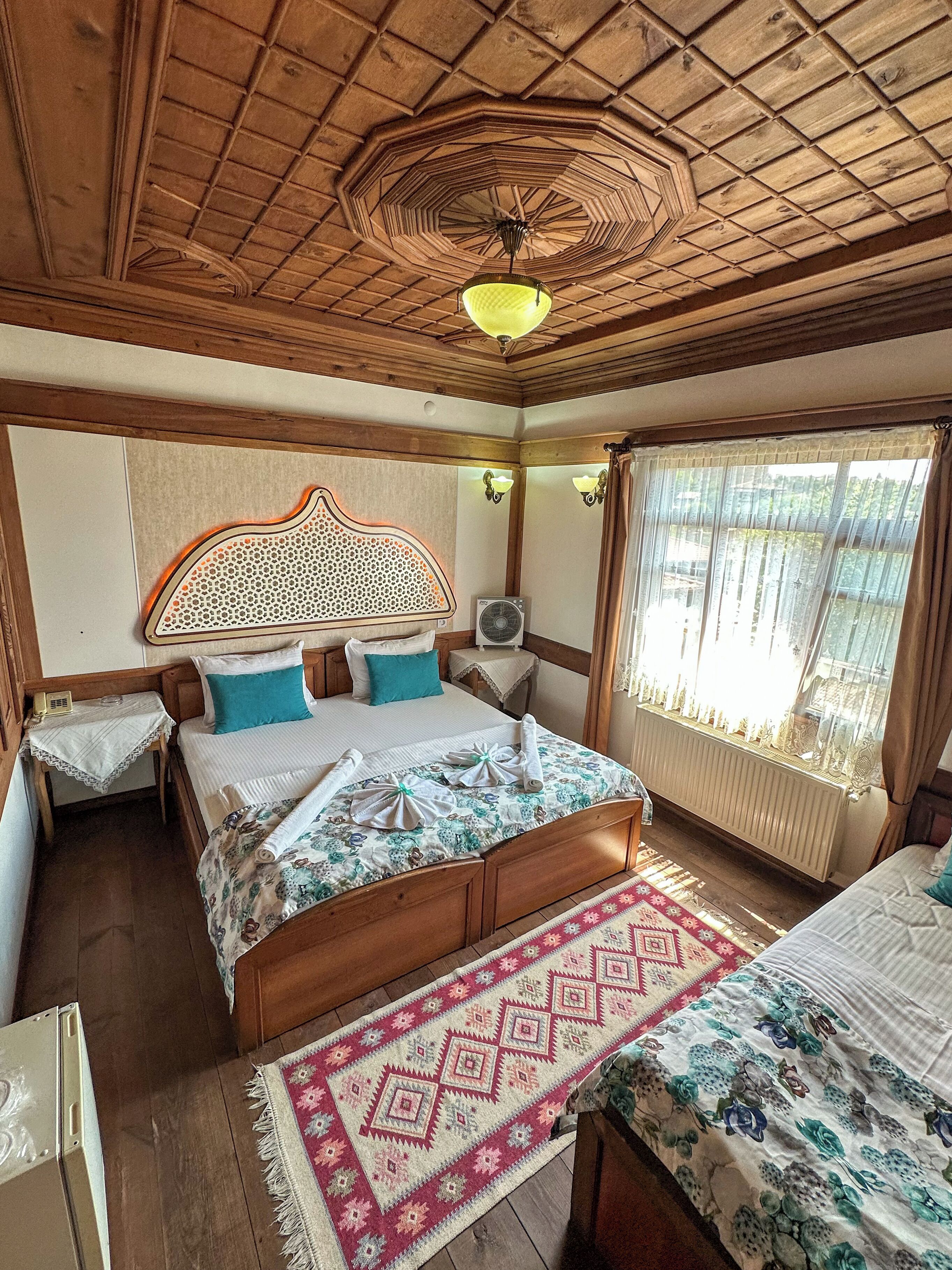 A bedroom with a wooden ceiling, a bed with floral bedding, a bedside table, a radiator, and a window with curtains.