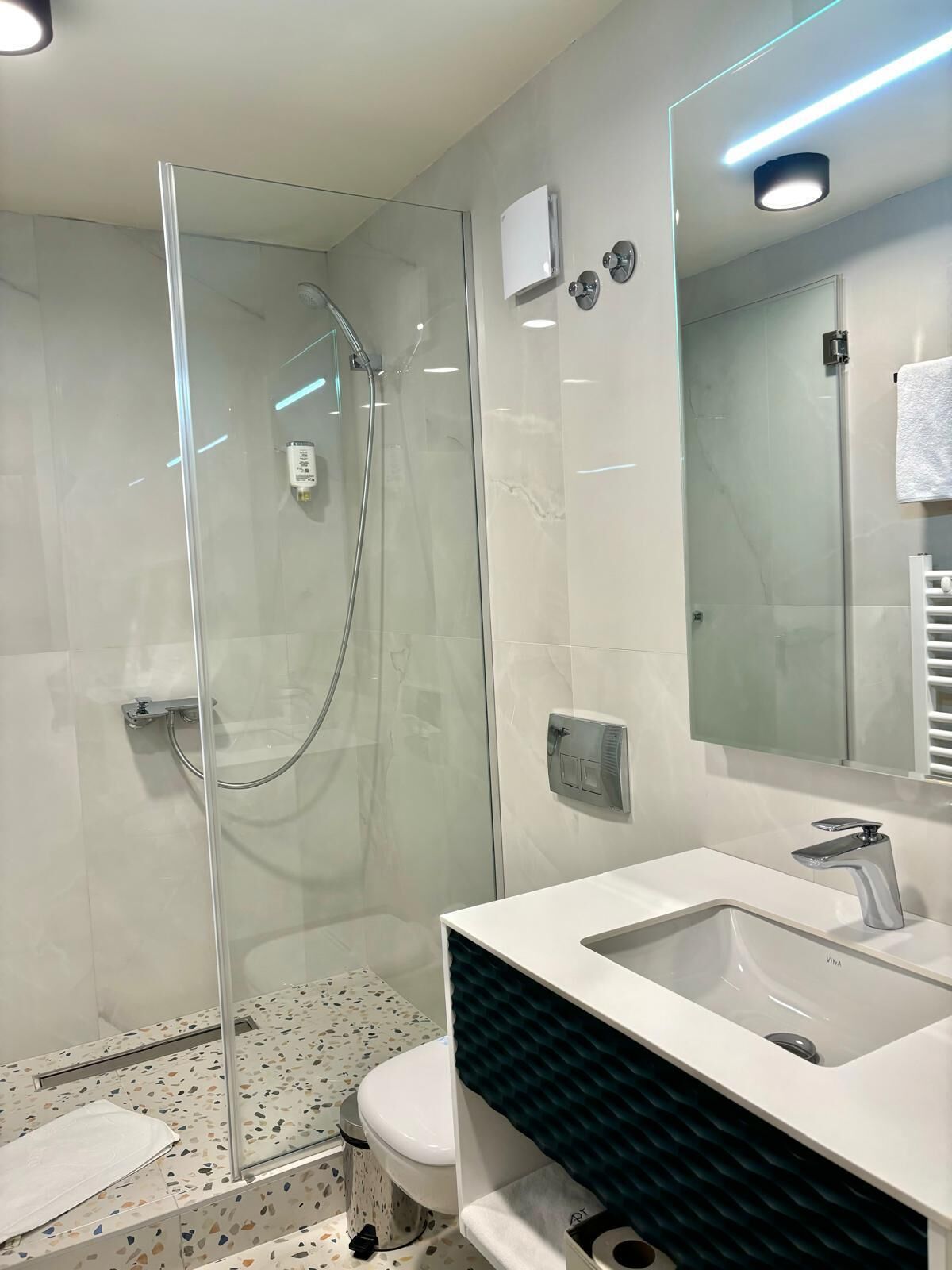 Un baño moderno con ducha de vidrio, un lavabo blanco con un solo grifo y un inodoro sobre un suelo con diseño.