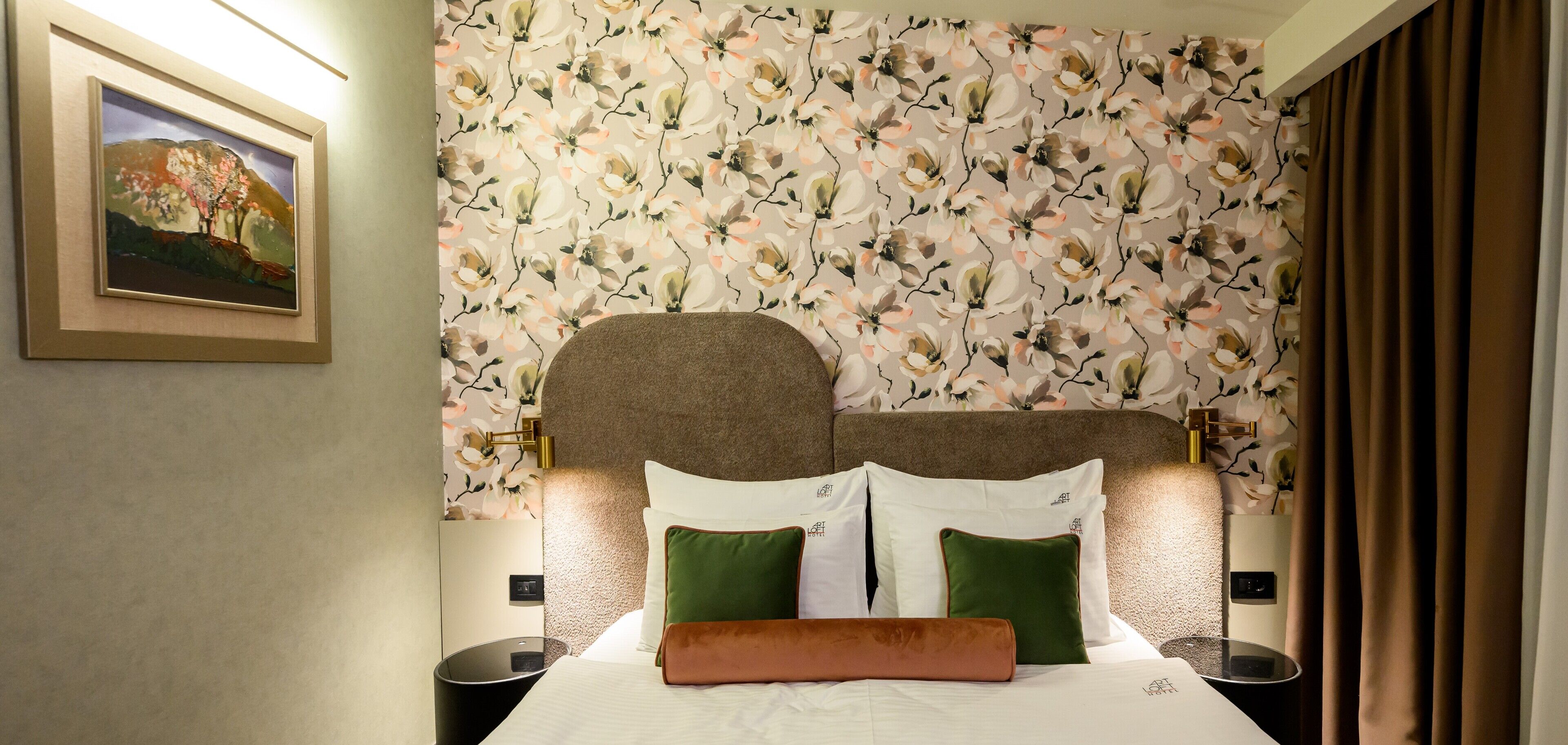 A hotel room with a floral wallpaper, a framed painting on the wall, and a bed with green and white pillows.