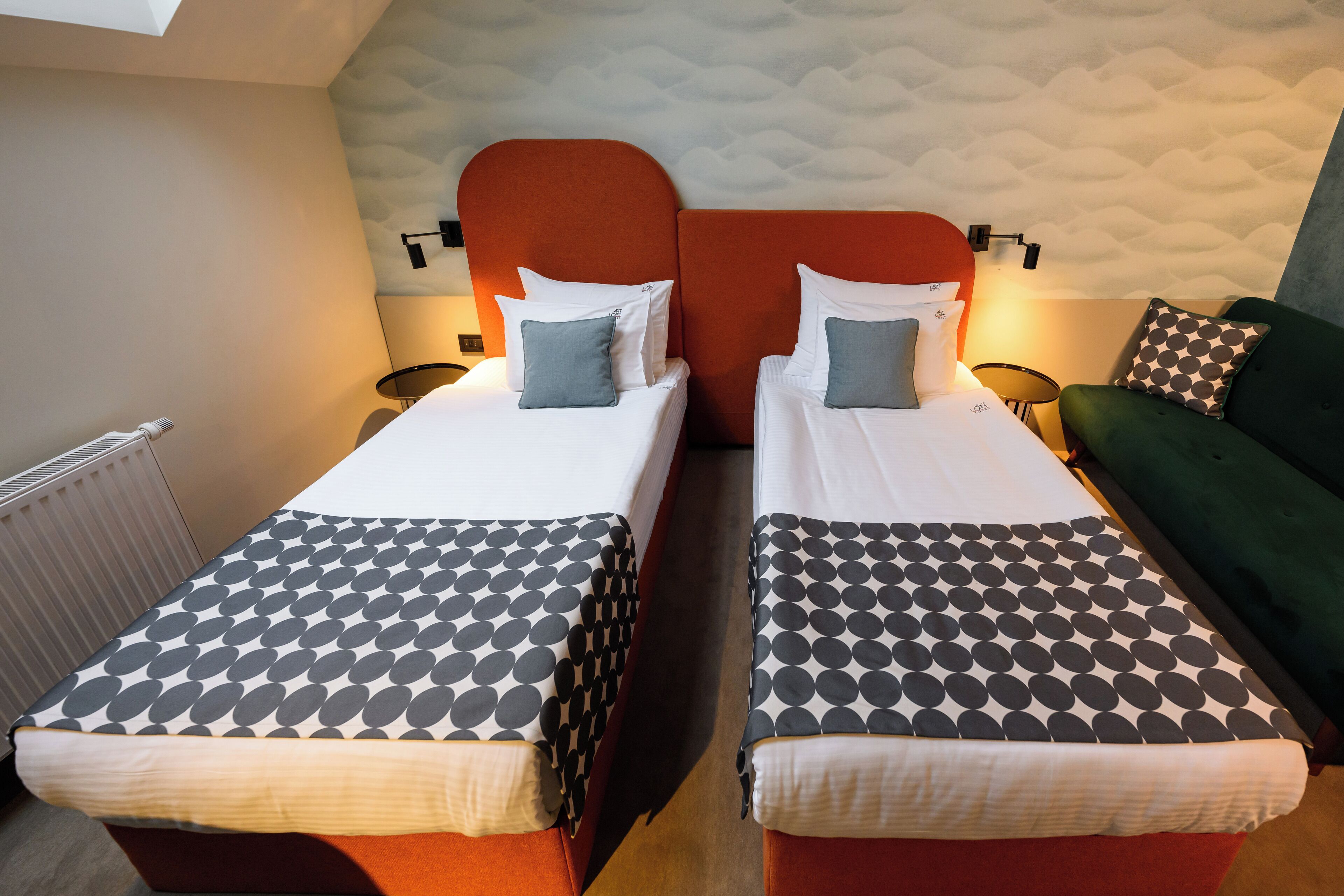 Dos camas individuales con ropa de cama estampada, un cabecero rojo y un sofá verde en una habitación con techo texturizado.