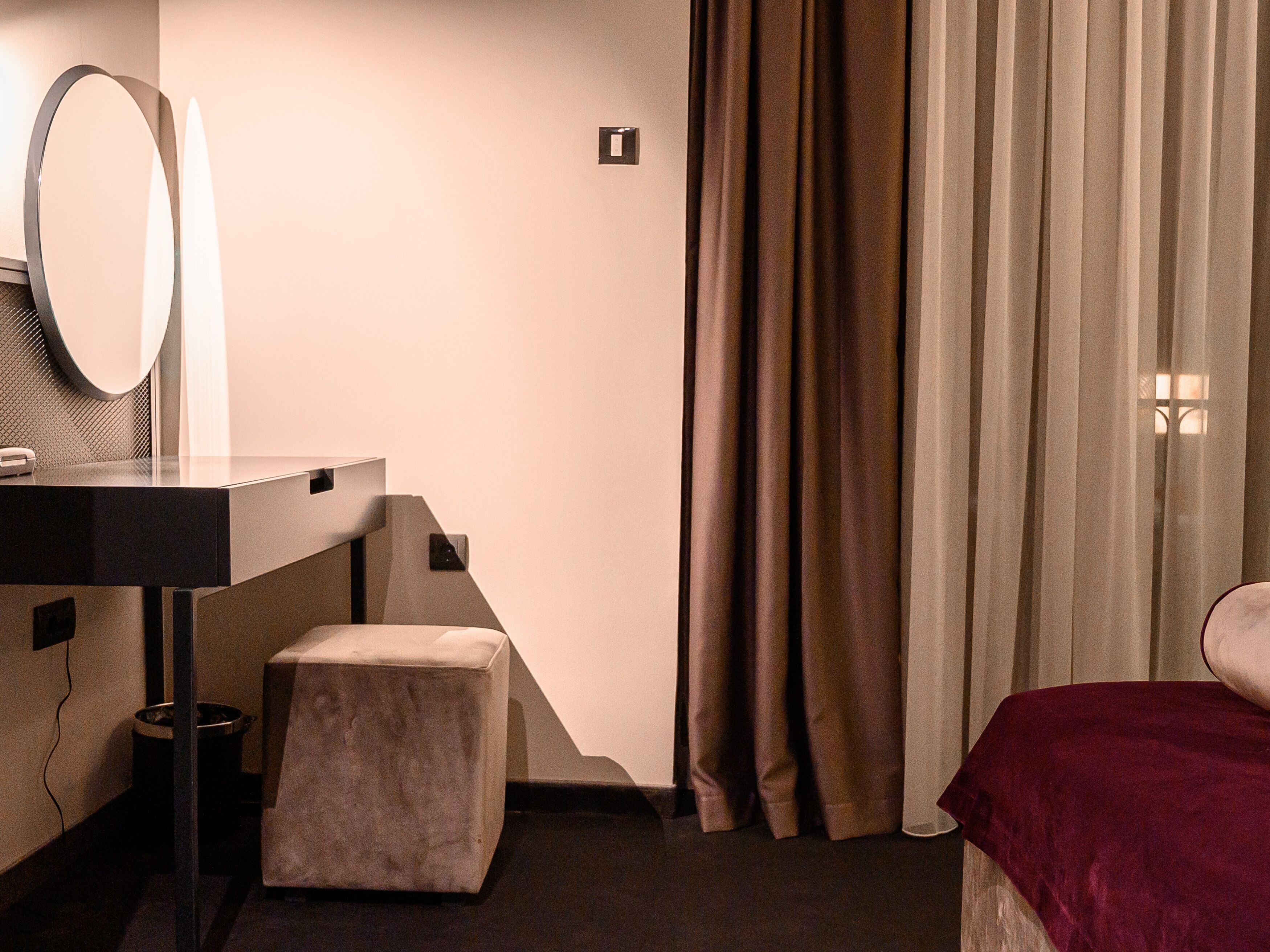 Een moderne hotelkamer met een donker houten bureau, een ronde spiegel, een klein krukje en een bed met een rode deken.