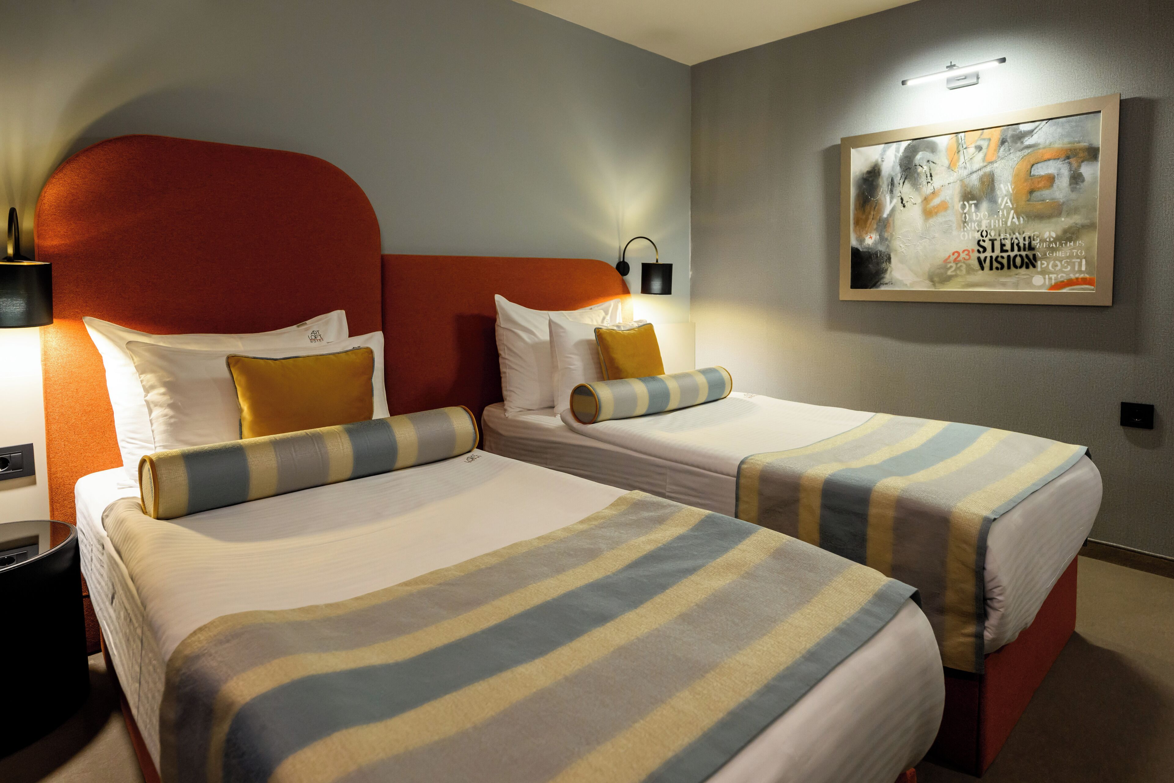 Una habitació d'hotel amb dos llits, cadascun amb roba de llit a ratlles, un panell de capçal vermell i una obra d'art penjada a la paret.
