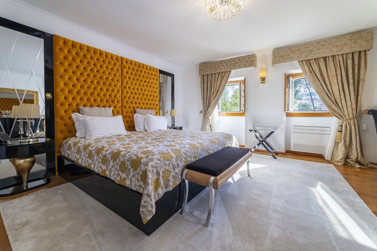 A bedroom with a tufted headboard, a large bed with a patterned bedspread, a bench at the foot of the bed, a desk with a lamp, and a large mirror.