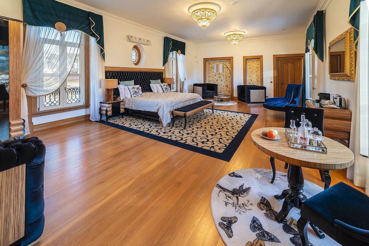 A spacious bedroom with a large bed, a sitting area with a table and chairs, and a view of the outside through a window with curtains.