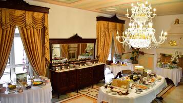 Uma sala de jantar formal com várias mesas dispostas para um buffet, um lustre grande e cortinas ornamentadas.