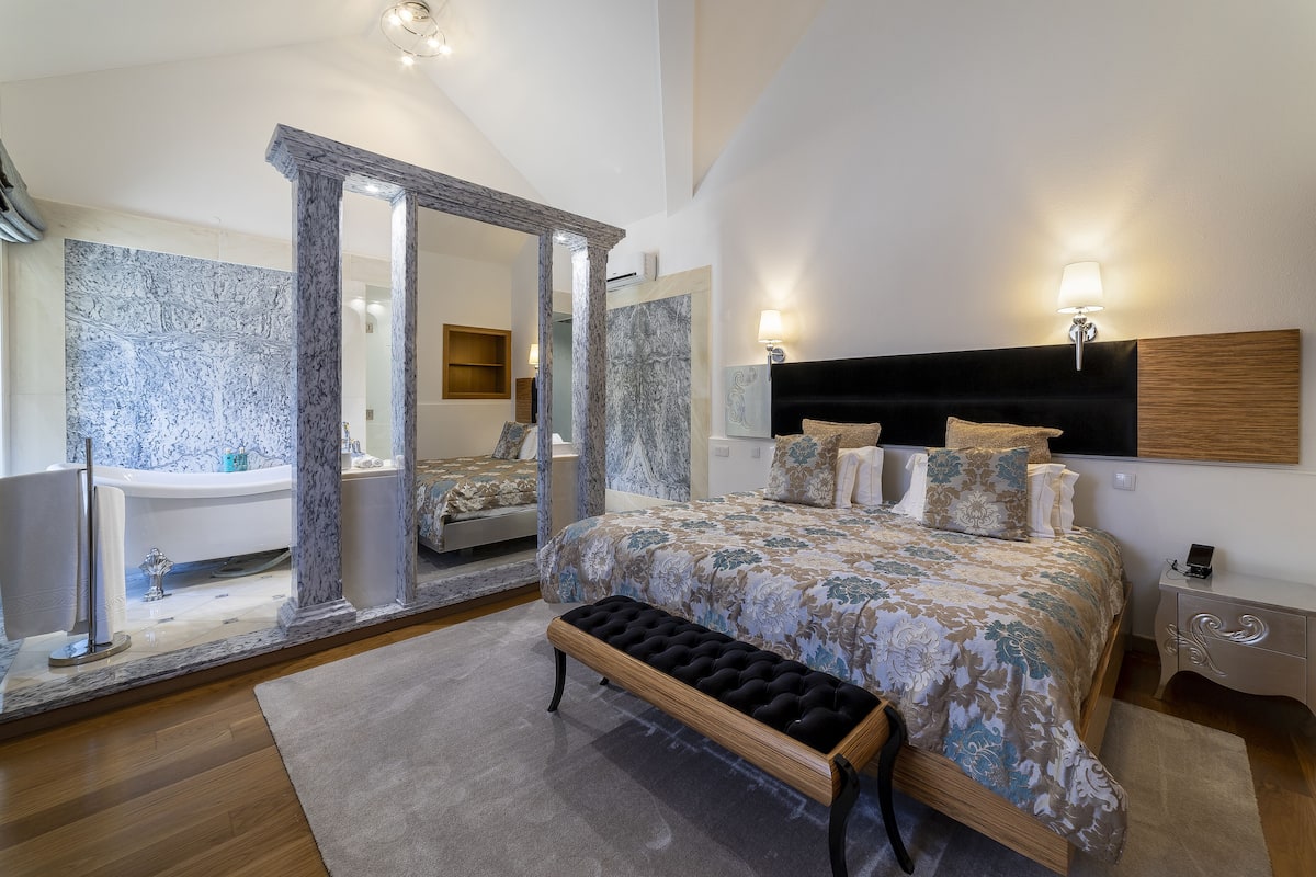 A bedroom with a large bed, a bench, a nightstand, a mirror, and a bathroom with a bathtub and a toilet.