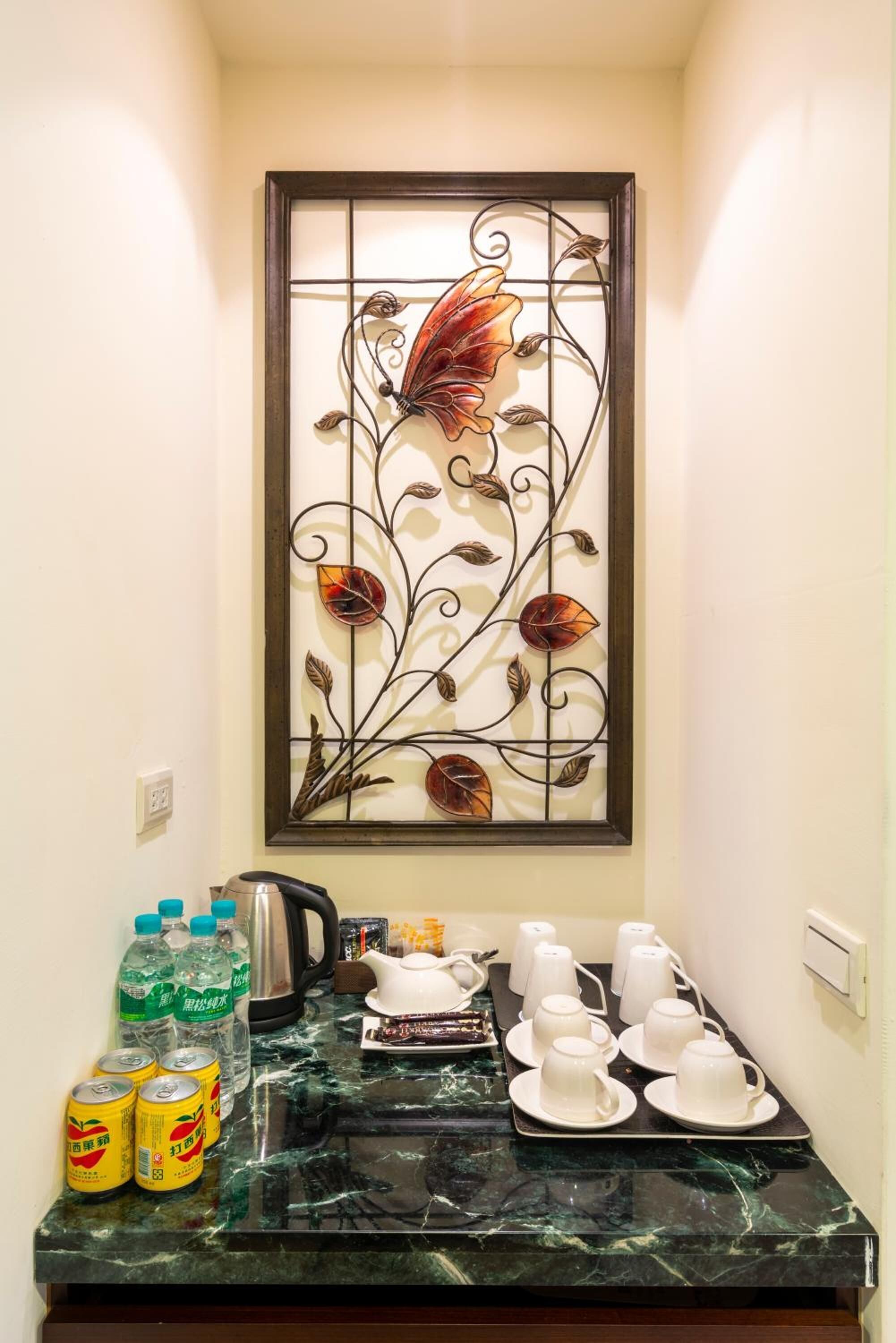 A hotel room with a decorative metal panel with floral designs, a marble countertop with tea service items, and bottled beverages.