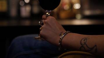 A hand holding a wine glass, seated on a yellow chair in a dimly lit room with a patterned wall in the background.