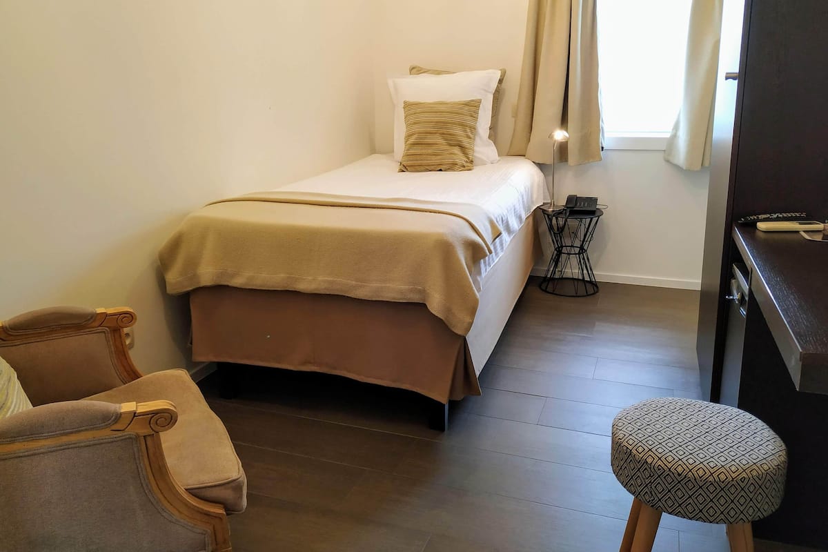A small, neatly arranged bedroom with a bed, bedside table, lamp, and a chair.