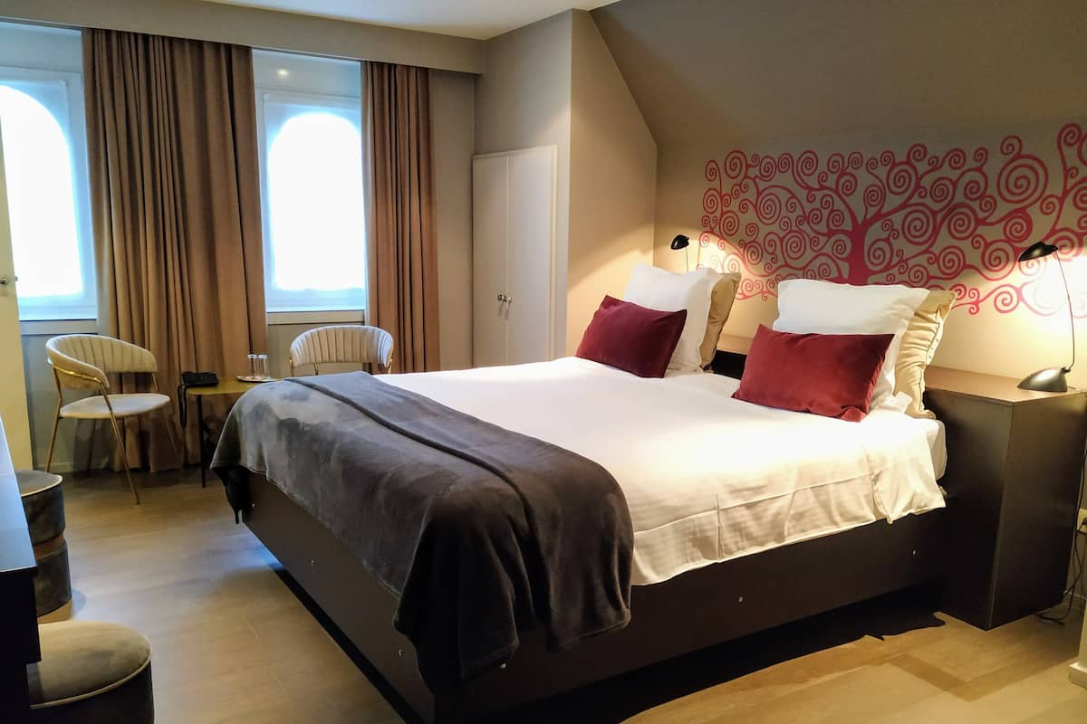 A modern hotel room with a large bed, a desk, two chairs, and a view of the outdoors through the window.