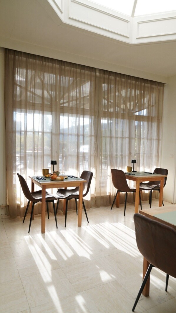 A dining area with two tables and chairs, large windows with sheer curtains, and a skylight.