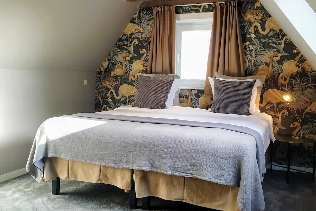 A bedroom with a sloped ceiling, a bed with a grey and beige bedspread, two pillows, and a nightstand with a lamp.