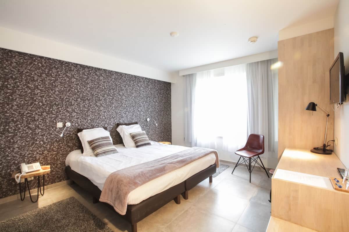 A modern hotel room with a large bed, a wooden desk, a chair, a TV, and a stone-textured wall.