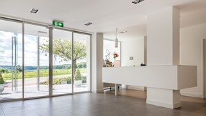 Un área de recepción moderna con puerta de cristal que da a un jardín, un mostrador de recepción blanco y un suelo limpio con baldosas.