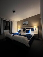 Quarto de hotel com uma cama grande, duas mesas de cabeceira, uma janela com cortinas, um quadro pendurado na parede e uma luminária no teto.