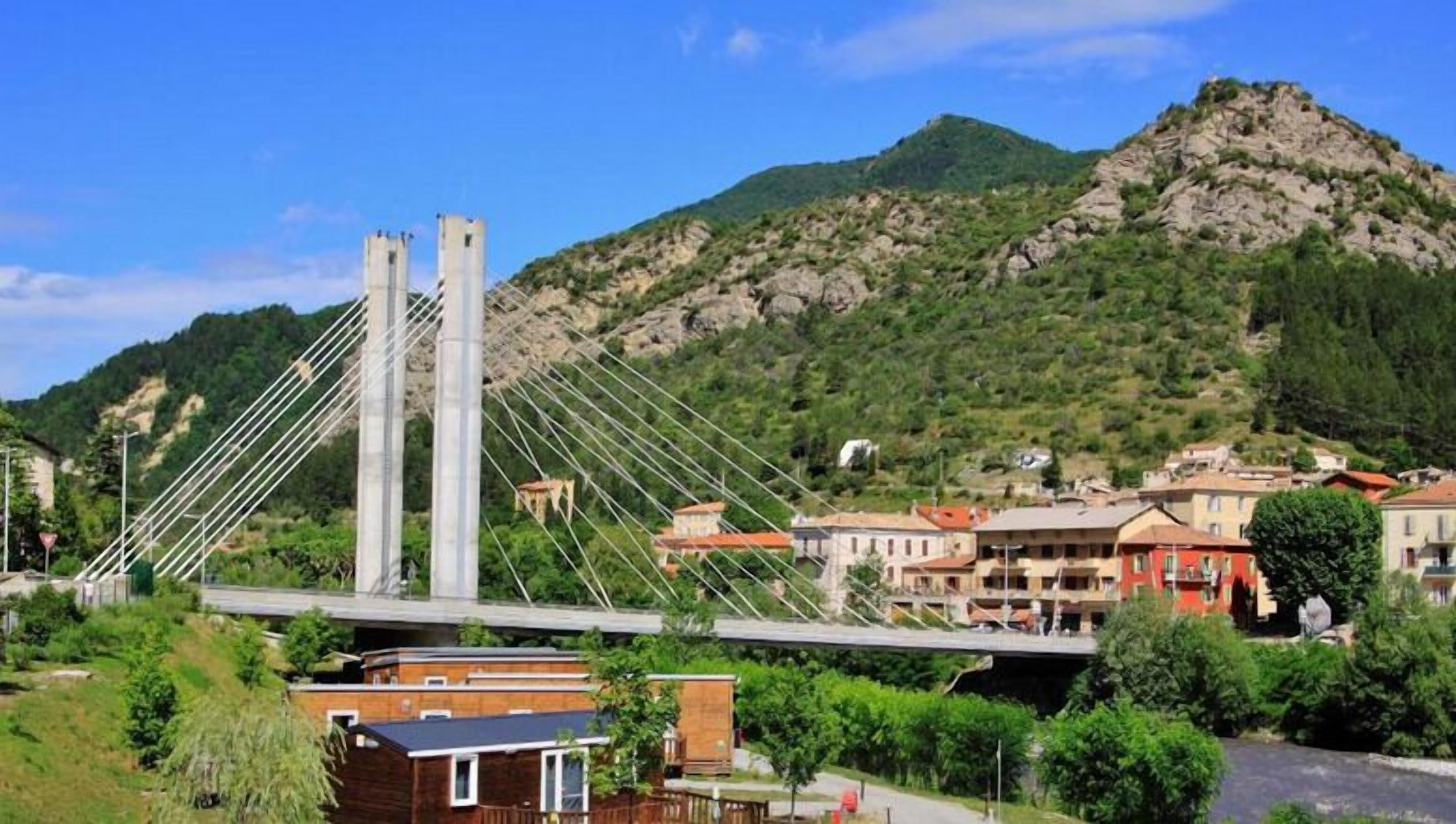 A cable-stayed bridge with a mountainous backdrop, a small building with a red roof, and a road leading to the bridge.