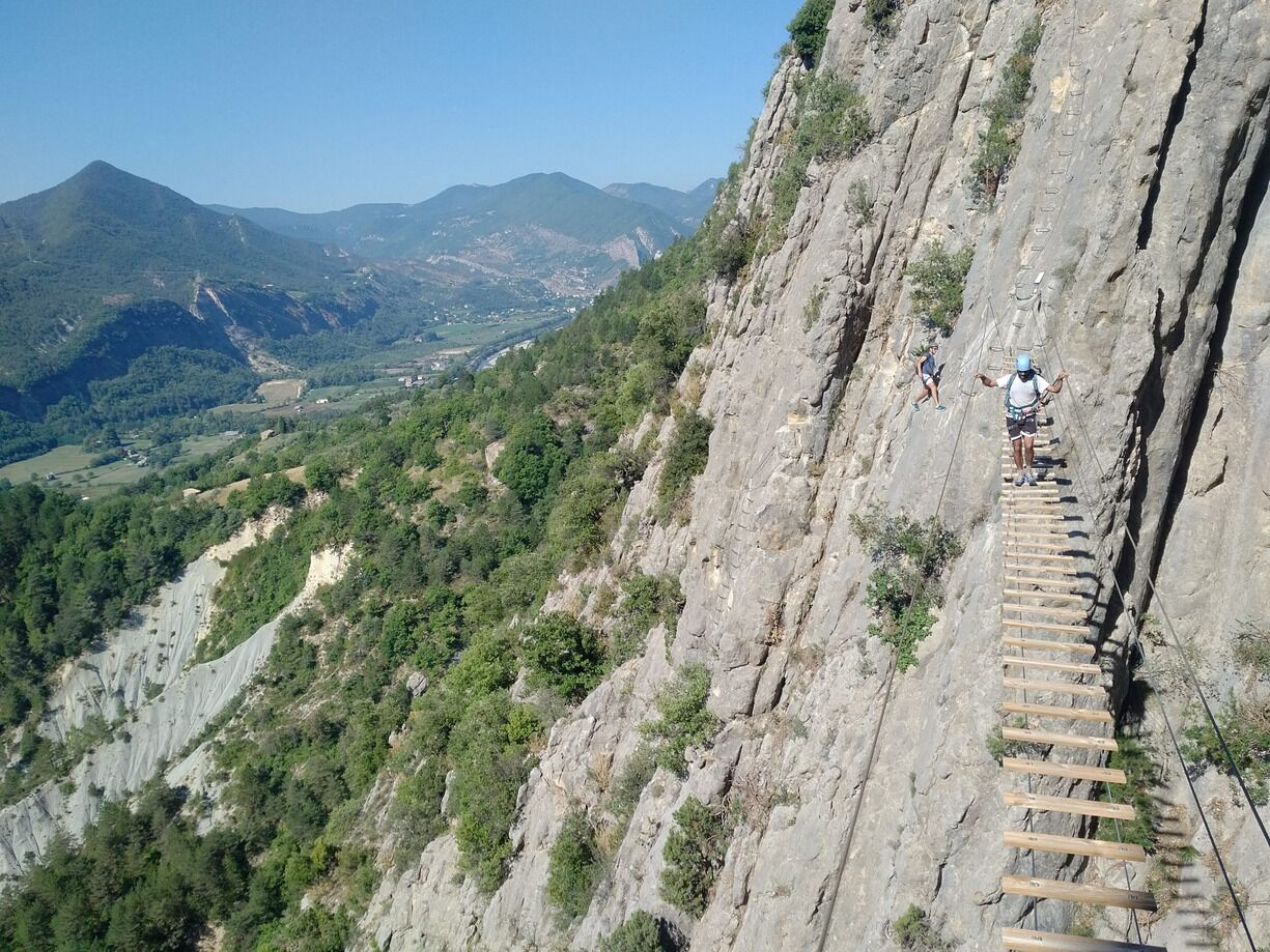 A wooden climbing route on a steep mountain cliff with a valley and distant mountains in the background.