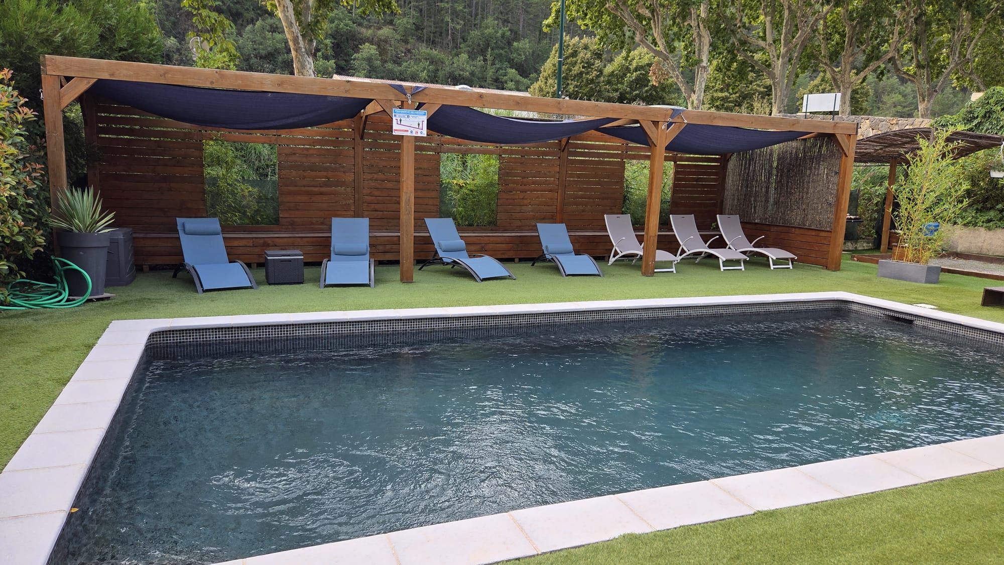 Piscina retangular com borda azulejada, rodeada por um deck de madeira. Várias espreguiçadeiras estão dispostas ao longo da borda da piscina.