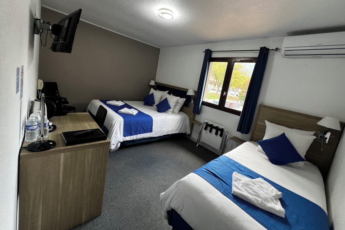 A hotel room with two beds, a desk, a chair, a TV mounted on the wall, and a window with curtains.