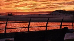Une vue sur le coucher de soleil, prise depuis un banc donnant sur une plage, avec une montagne lointaine et une jetée.