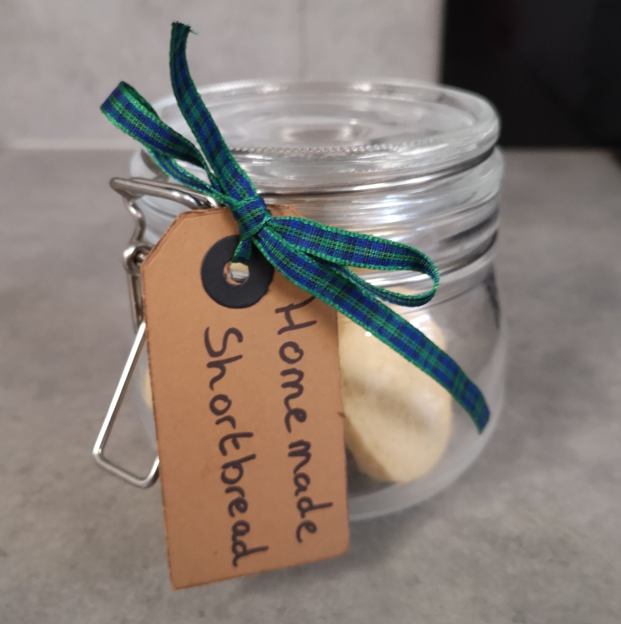 A glass jar with a metal clasp lid, containing a loaf of bread, tied with a green and blue ribbon. A tag attached to the ribbon reads "Homemade Shortbread."
