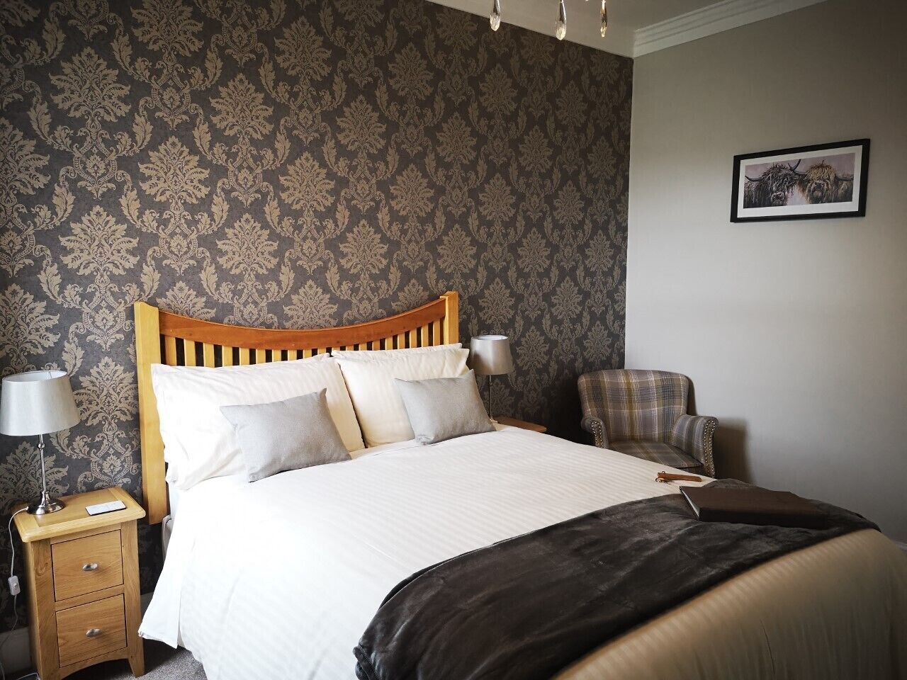 A neatly made bed with a wooden headboard, a plaid armchair, and a bedside table with a lamp.