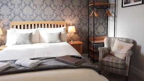 Un lit bien fait, agrémenté d’oreillers, une table de chevet avec une lampe, un fauteuil à motifs et un tableau encadré au mur.