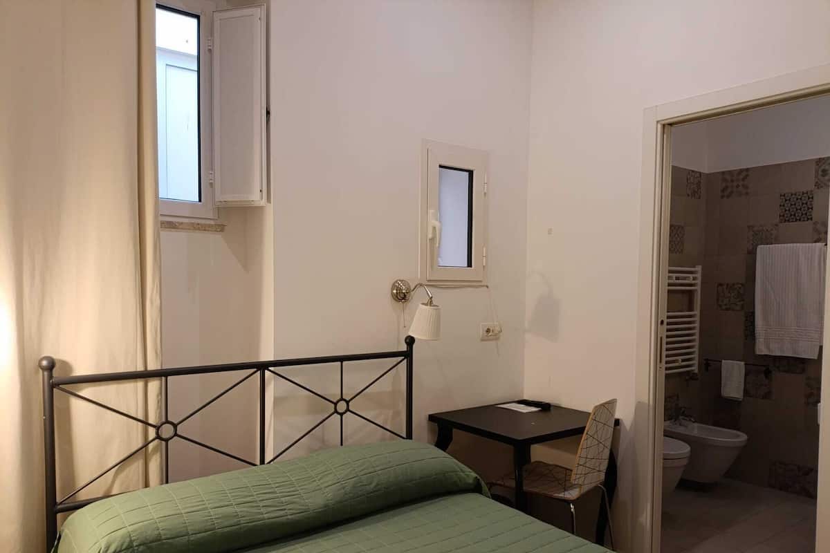 Un lit simple avec un cadre en métal et une literie verte, une petite table avec une chaise, une lampe fixée au mur et une fenêtre avec un rideau.