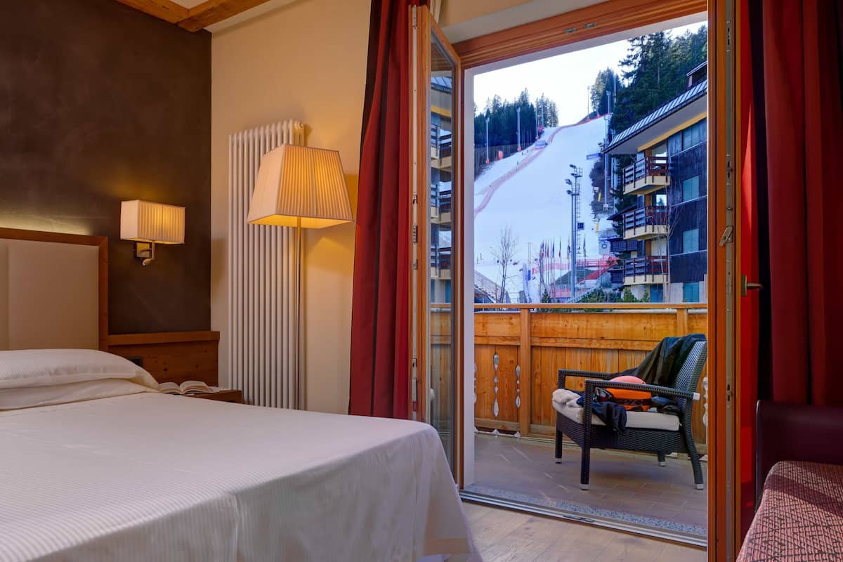 A hotel room with a bed, a nightstand, a lamp, a window with curtains, and a view of a ski slope.