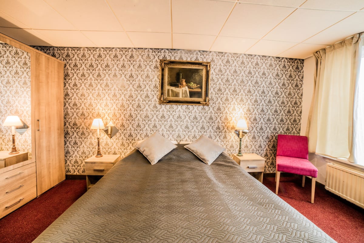A hotel room with a large bed, two bedside tables with lamps, a chair, a wardrobe, and a painting on the wall.