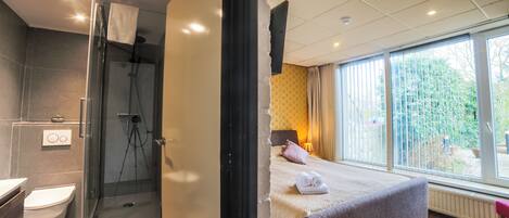 A modern hotel room with a shower, toilet, and a bed with a view of the outdoors.