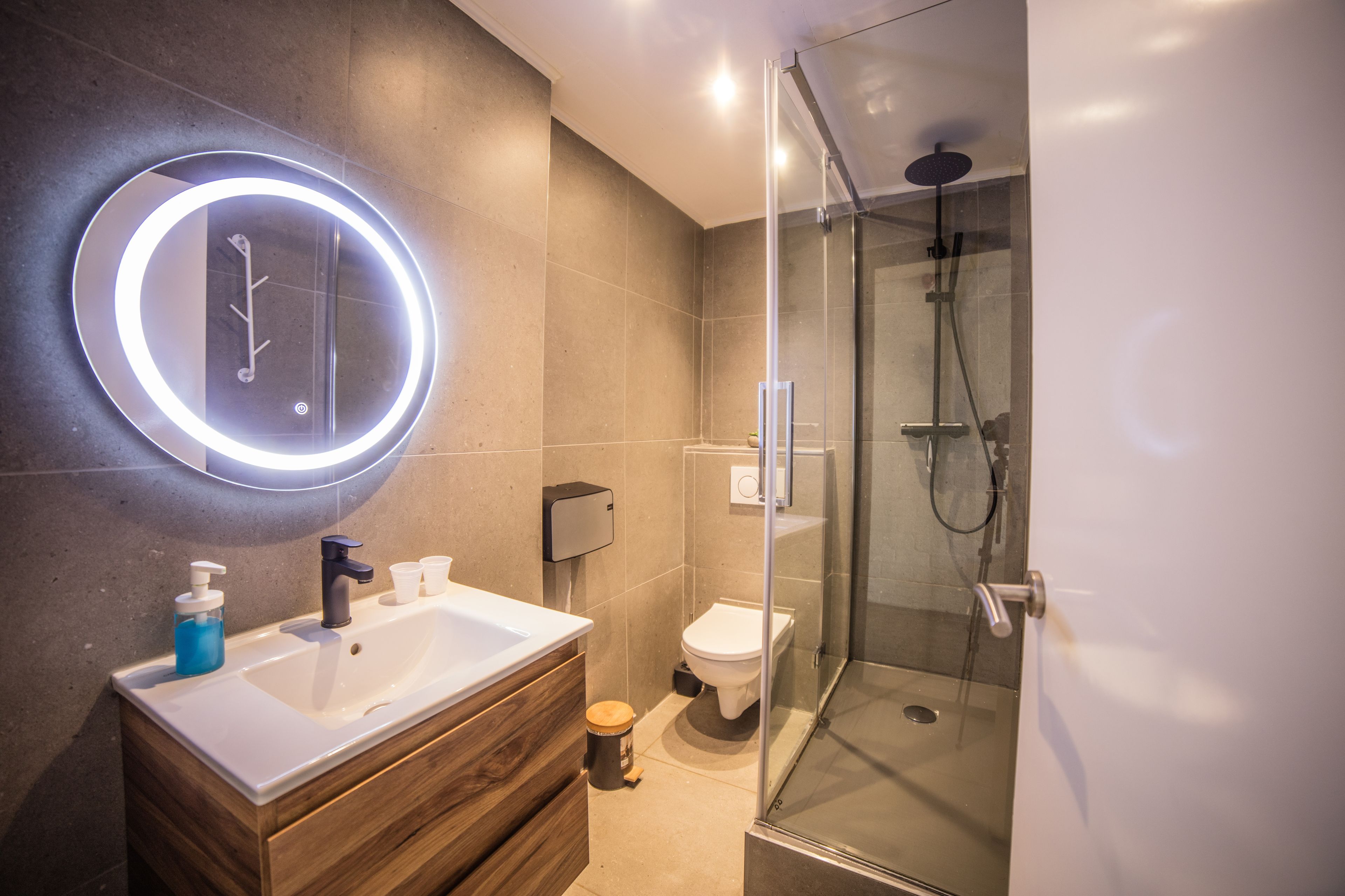 Un baño moderno con ducha envuelta en vidrio, espejo redondo con iluminación, lavabo blanco y tocador de madera.