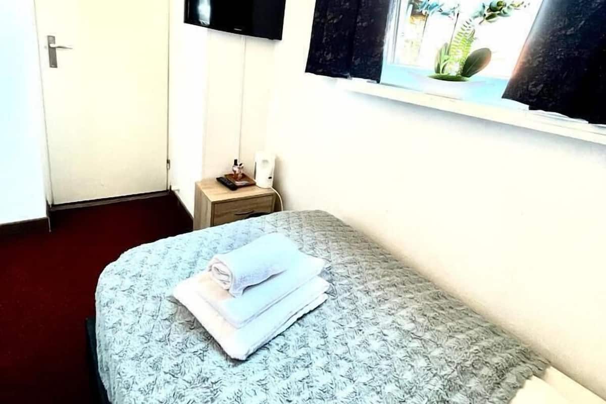 A hotel room with a bed, a TV mounted on the wall, a bedside table with a lamp, and a window with curtains.
