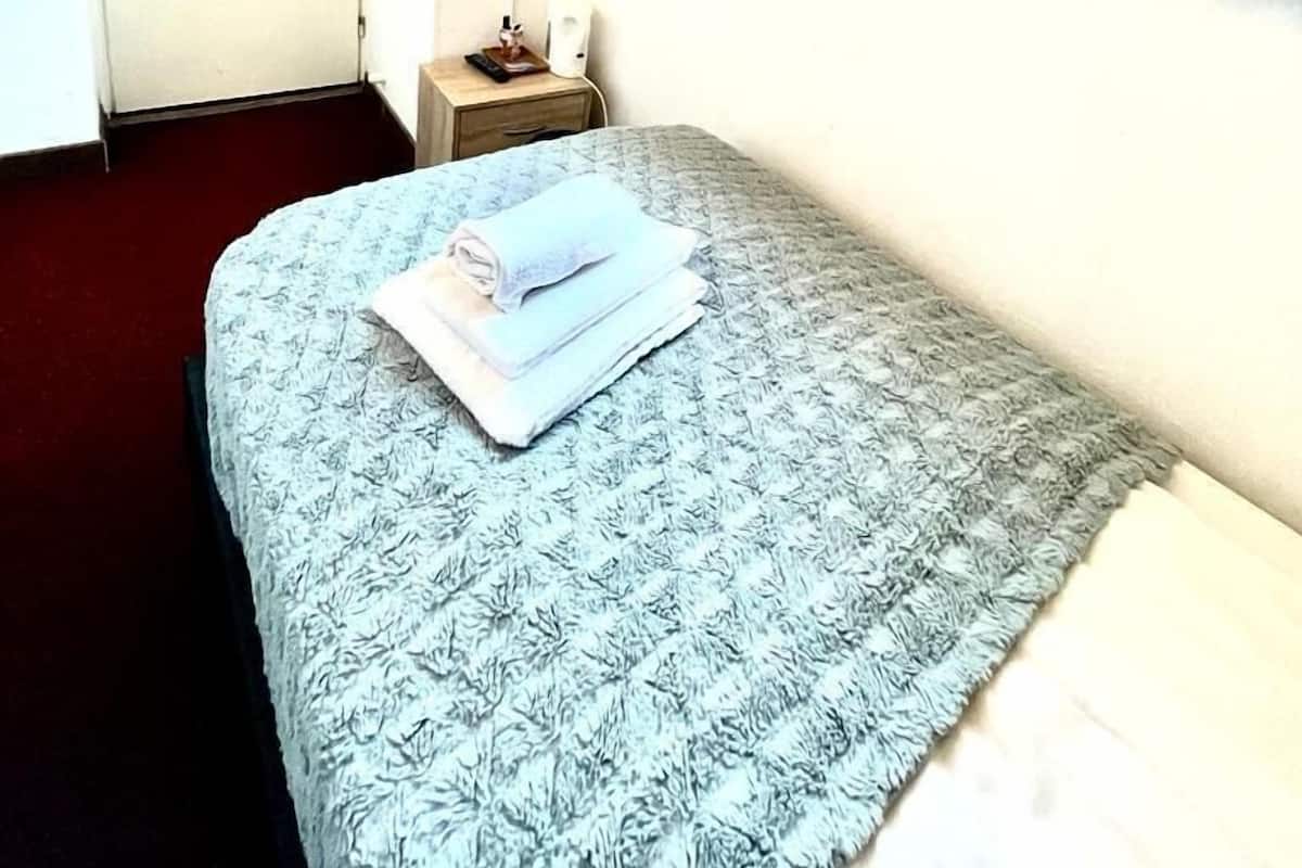 A small, simple room with a bed, a TV mounted on the wall, a bedside table, and a window with curtains.